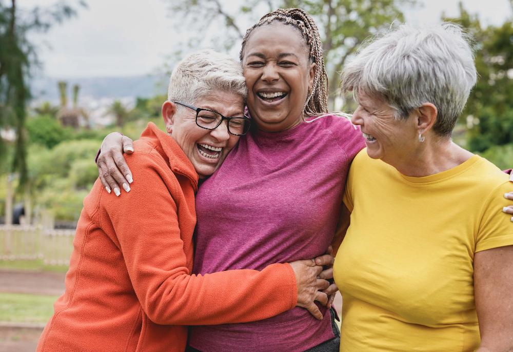Three diverse older women laughing and embracing outdoors, showing joy and connection