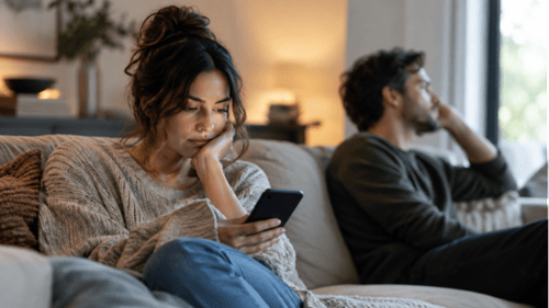 Woman looking at her phone while her partner sits turned away on the couch, reflecting emotional dis